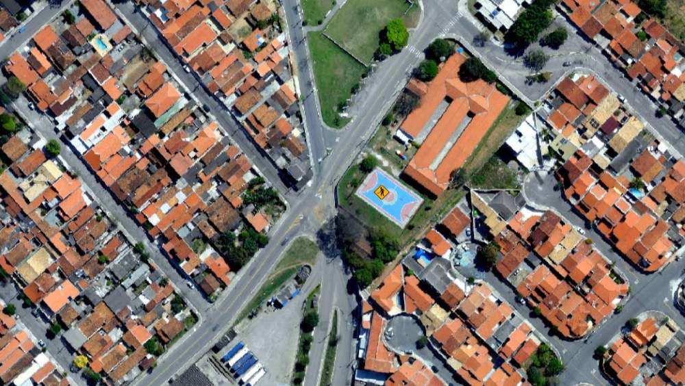 A imagem mostra um exemplo claro dos resultados obtidos por meio de aerolevantamento: uma captura aérea de alta resolução que permite visualizar com nitidez a malha urbana, incluindo ruas, quadras, construções e equipamentos públicos, como a quadra esportiva no centro da imagem. 